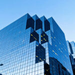 Modern glass office building with distinctive vertical dark window columns against a clear blue sky, surrounded by other high-rise buildings in an urban setting.