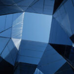 Upward view of angular glass and metal architectural surfaces with overlapping geometric planes in shades of blue, reflecting clear sky between modern building structures.