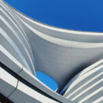 Upward view of a modern white building facade with curved architectural lines and horizontal striping, framing clear blue sky in the center opening.