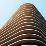 Upward view of a curved modern building with stacked horizontal balconies in brown and tan tones against a clear blue sky.