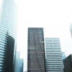 Upward view of multiple modern glass and steel skyscrapers with varying architectural styles and window patterns against an overcast sky in an urban downtown setting.