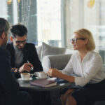 Three business professionals in casual meeting attire having a discussion in a modern high-rise office lounge with floor-to-ceiling windows overlooking a city skyline, with coffee cups and notebooks on the table.
