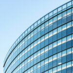 Curved modern glass and steel building facade with repetitive rows of rectangular windows in blue and white tones against a clear light blue sky.