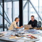 Two professionals collaborating at a table in a high-rise office with floor-to-ceiling windows showing city buildings, surrounded by design materials, color swatches, and a laptop with a cityscape view in the background.